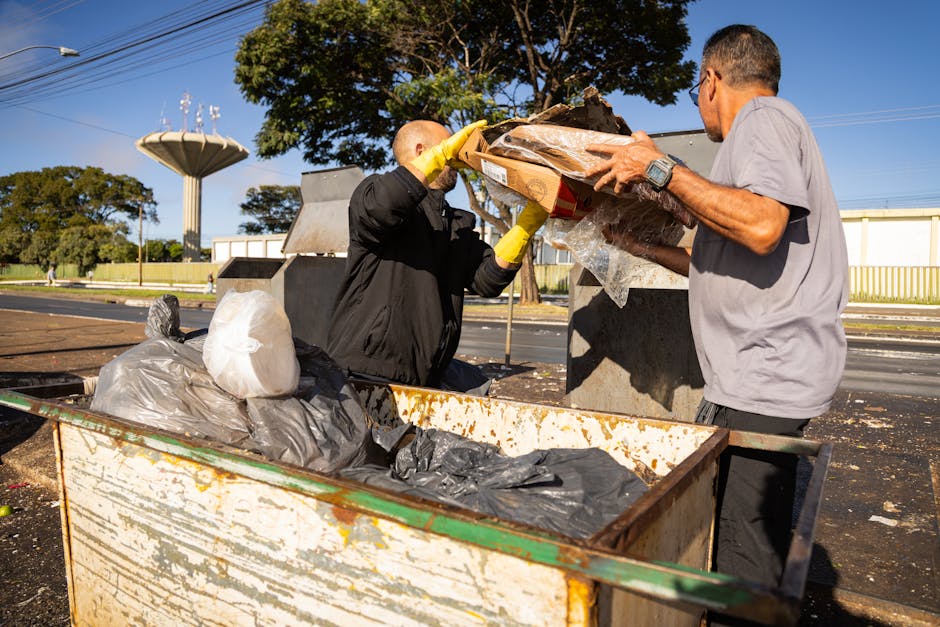 Two men are engaged in a rubbish collection process outdoors next to a large, rusty metal dumpster. One man, wearing a black jacket with yellow gloves, is lifting a cardboard box filled with plastic-wrapped items and debris over the open top of the dumpster. The second man, dressed in a grey t-shirt and dark trousers, is helping him by holding the box steady. The dumpster contains various black and grey rubbish bags, some of which are partially open, revealing plastic waste. Behind them, there are clear blue skies and trees, with a large water tower visible in the background. The scene is set on a paved area, adjacent to a road with some distant industrial or commercial buildings. The lighting is natural and bright, highlighting the ongoing waste disposal activity as part of a home relocation or clearance process, relevant to house removals and furniture transport services offered by Man with Van Cheam.