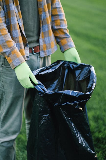 A person wearing a green long-sleeved shirt, a plaid jacket, and light-colored trousers is standing outdoors on a grassy area, holding open a black trash bag lined with a shiny silver interior. The individual's hands are covered with light green gloves, and they appear to be preparing to dispose of or transport waste. In the background, there is a lush, well-maintained lawn, suggesting a garden or outdoor yard setting. The scene is well-lit with natural daylight, emphasizing the process of waste collection or cleanup as part of house removals or home relocation services, such as those offered by Man with Van Cheam. The image captures the practical aspects of packing, loading, or disposing of items during a relocation or clearance process, focusing on the careful handling of waste materials, possibly furniture packaging or household rubbish. This step could relate to efficient moving logistics, including packing and loading in preparation for transport to a new residence.