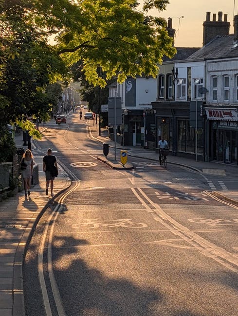 A quiet street scene captured during late afternoon or early evening, with a large tree casting shadows over the road. Pedestrians are walking along the pavement, including two women in casual clothing walking away from the camera on the left side, and a cyclist riding on the right side near a row of shops. The street features tramway tracks embedded in the asphalt, with some vehicles visible further down the road. On the right, there are traditional shopfronts, one of which has a sign partially visible, and a person standing outside. The scene is illuminated by warm, golden sunlight filtering through the leaves, creating a relaxed atmosphere characteristic of a neighbourhood suitable for house removals and local moving services offered by Man with Van Cheam.