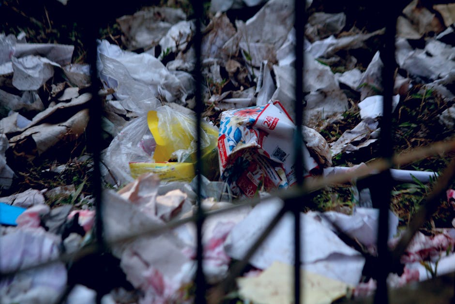 A close-up view of a person's hand holding a blue plastic rubbish bag filled with waste, the bag appears tightly gathered at the top with the hand gripping it securely. The background is neutral and o