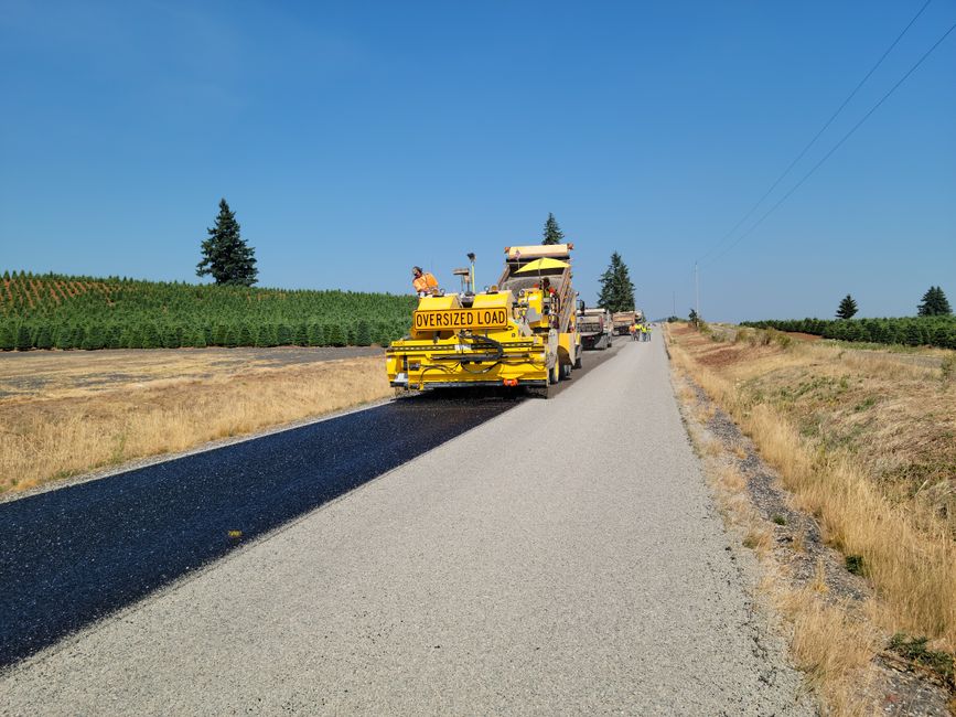 A road construction vehicle with bright yellow colouring and an 'OVERSIZED LOAD' sign at the rear is paving a section of the road with fresh black asphalt, while workers in safety vests operate machin
