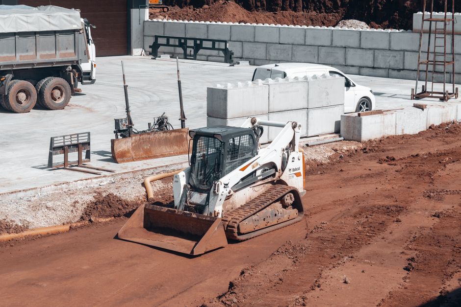 A road construction vehicle with bright yellow colouring and an 'OVERSIZED LOAD' sign at the rear is paving a section of the road with fresh black asphalt, while workers in safety vests operate machin