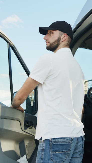 A man wearing a black cap, white t-shirt, and blue jeans is standing beside a moving van with its side door open. He appears to be engaged in loading or unloading items, with his arms resting on the v
