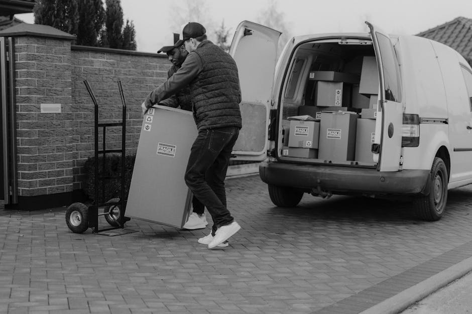 A man wearing a black cap, white t-shirt, and blue jeans is standing beside a moving van with its side door open. He appears to be engaged in loading or unloading items, with his arms resting on the v