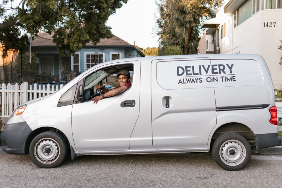 A silver delivery van parked on a residential street with the words 'DELIVERY ALWAYS ON TIME' printed on its side. Inside the cab, a smiling driver is visible, wearing a maroon shirt and glasses, with one arm resting on the open window frame as he looks towards the camera. The van is positioned near a white picket fence and a sidewalk, with a blue house featuring white window frames and a dark roof in the background, along with trees and greenery. The scene suggests the loading or unloading process associated with home relocation or furniture transport, as part of a professional removals service. The environment is well-lit by natural daylight, highlighting the vehicle, the driver, and the surrounding residential setting, aligning with the context of house removals and moving logistics.