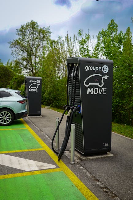 A view of two electric vehicle charging stations positioned on a paved parking area adjacent to a house removal site, with a grey car parked nearby. The larger charging unit in the foreground displays the logo 'groupe e MOVE' and features an electric plug graphic, with black charging cables hanging and connected to the unit. The smaller charging station in the background also bears the same branding and logo. The scene is set outdoors, surrounded by lush green trees and a partly cloudy sky, indicating daytime. The parking spaces are marked with green and yellow lines, designating areas for electric vehicle recharge during a home relocation or furniture transport process, which may be part of the logistics managed by Man with Van Cheam, a professional removals company.