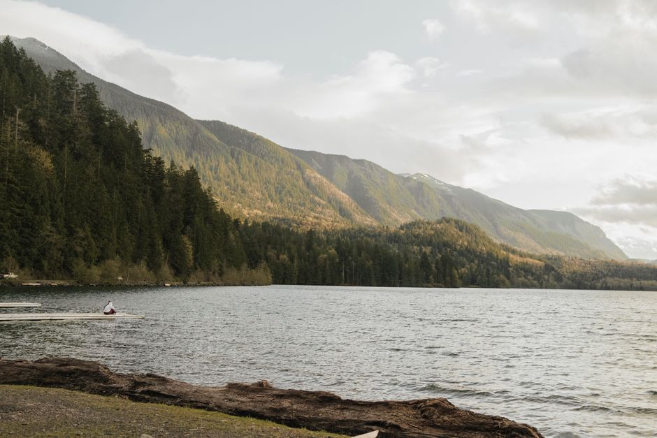This image shows a tranquil lakeside scene with a calm body of water surrounded by lush green forested hills and mountains in the background. The sky is partially overcast with soft, diffused lighting illuminating the landscape. On the left side, a person dressed in casual outdoor clothing is sitting in a small rowing boat near the water's edge, which is lined with rocks and logs. The shoreline features scattered natural debris and vegetation, creating a peaceful, natural environment typical of a remote outdoor location. Although the scene is serene and focused on nature, it subtly emphasizes the importance of logistics and transport in remote settings, reflecting the context of house relocations or outdoor moving activities that Man with Van Cheam might facilitate in rural or scenic areas.