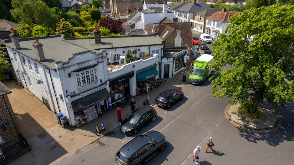 An aerial view of a busy street corner in Cheam, showing a small white commercial building with signage and outdoor display areas, including flowers and potted plants. Several vehicles, such as black and grey cars and a green van, are parked along the roadside, while a green vehicle is being loaded or unloaded nearby. Two pedestrians, dressed casually, walk across the crosswalk near the building. To the right, a large leafy tree provides shade over the pavement and part of the parking area. The street is lined with residential houses and greenery, indicating a suburban setting suitable for house removals and furniture transport services. This scene captures the context of relocation logistics, with activity around loading and unloading within a neighbourhood environment, and the interaction between commercial premises and vehicle movement supported by Man with Van Cheam for efficient home relocation and moving services.